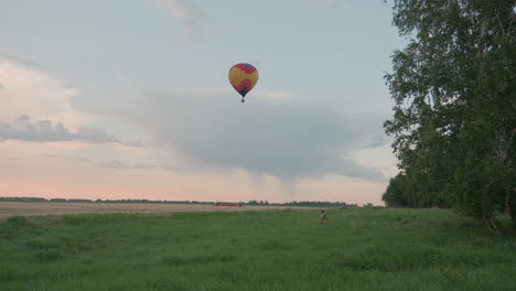 male crew member walking through tall grass holding tether rope guiding ascending hot air balloon into pastel sky over open field at dusk preflight launch scene