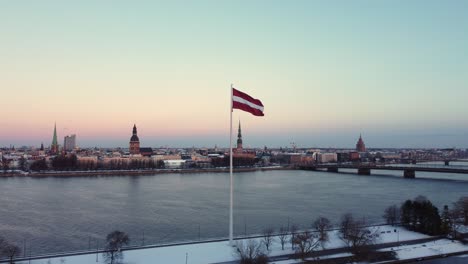 latvian flag waving by daugava river with cityscape, winter snowy city, aerial