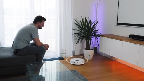 el hombre se sienta en un sofá en la sala de estar y activa su robot aspirador a través del control de voz del teléfono inteligente.