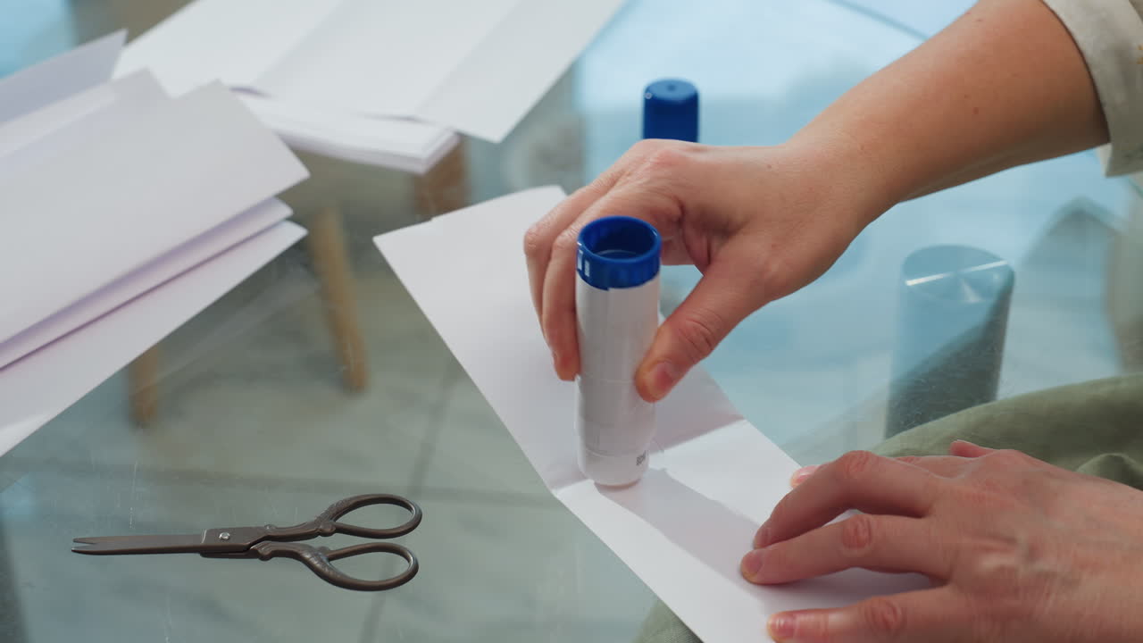 Woman hand applying liquid glue between paper and folding it, scissors and paper visible on table, showcasing crafting activity and attention to detail in a cozy home setting
