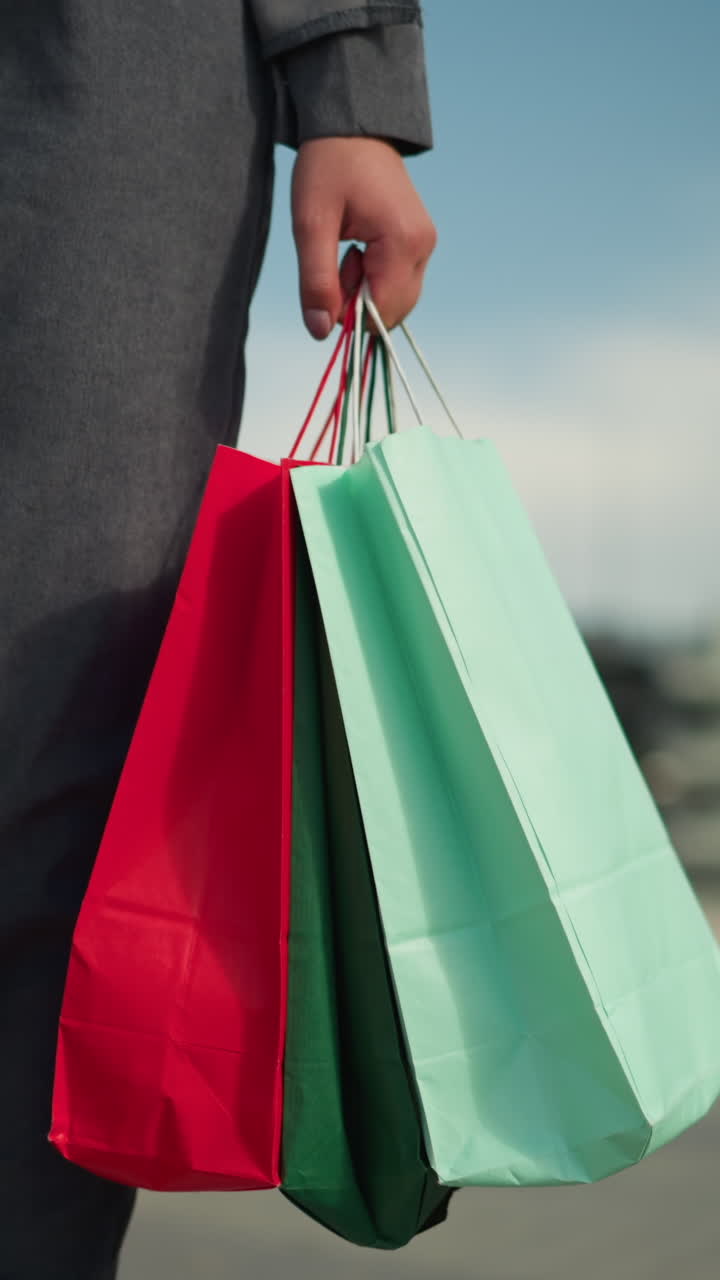 vista de la pierna de una dama con ropa gris sosteniendo tres bolsas de compras coloridas en su mano derecha mientras camina cerca de una valla decorativa, con un fondo borroso de elementos urbanos y cielos despejados