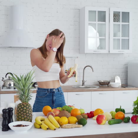 Attractive model eating banana. Healthy fruit and vegetables on the table. Cheerful woman eats fruit in the modern light kitchen.