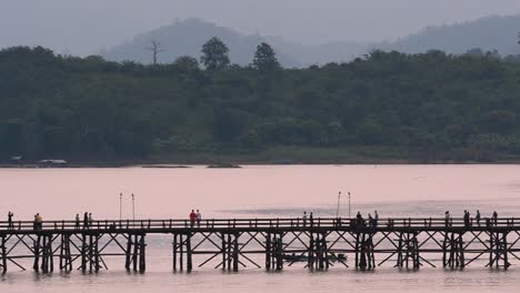 Mon-Bridge-and-a-longboat-followed-speeding-behind-the-structure-while-people-are-walking-on-the-bridge,-silhouetting-as-it-was-getting-dark,-in-slow-motion