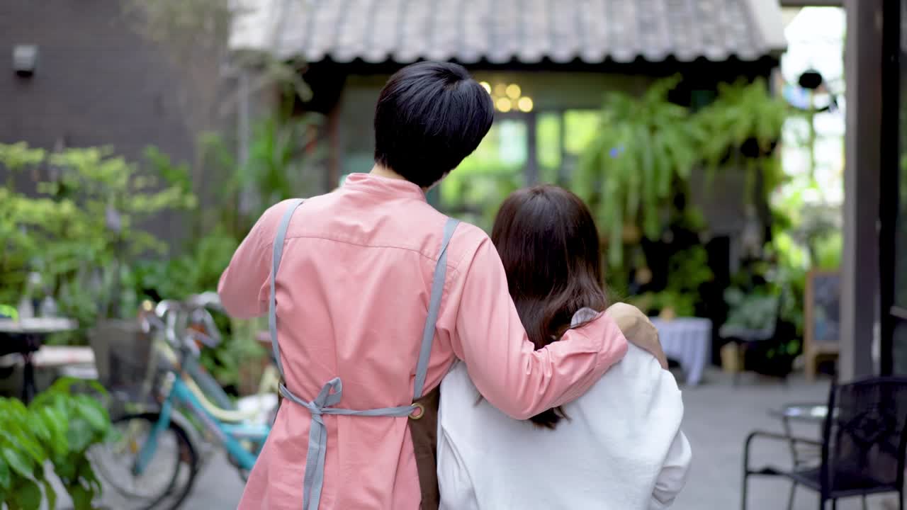 매력적인 젊은 아시아 부부는 새로운 레스토랑의 개막을 축하하기 위해 기했습니다. 우리는 문을 열고 있습니다. 앞 문에 표지판이 웃으며 새로운 카페에 고객을 환영합니다.