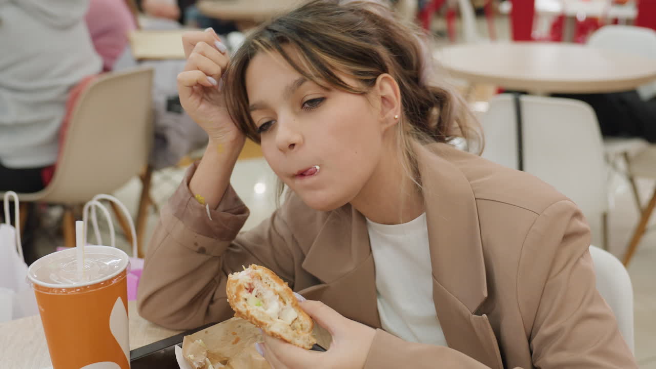 Белый пассажир ест бургер с кунжутом в фуд-корте торгового центра, рядом большая оранжевая чашка, оживленная посадка, сосредоточенно жует, повседневный блейзер, обеденный перерыв, крошки и удовольствие, момент перекуса на ходу