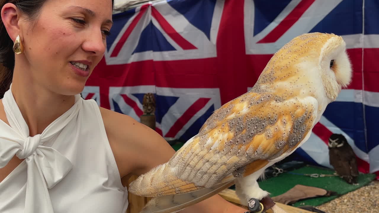 Close-up of a woman in a white dress holding a barn owl beside British flags. The scene is warm and inviting with intricate details on the bird's feathers