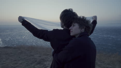 man holding fabric by frozen lake as romantic girlfriend embracing him