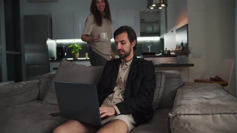 Happy-brunette-man-with-stubble-in-a-black-jacket-works-at-his-gray-laptop-while-his-brunette-girlfriend-in-a-beige-T-shirt-brings-him-tea-on-the-sofa-in-a-modern-studio-apartment-in-the-evening