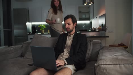 feliz hombre moreno con rastro en una chaqueta negra trabaja en su portátil gris mientras su novia morena en una camiseta beige le trae té en el sofá en un apartamento estudio moderno por la noche