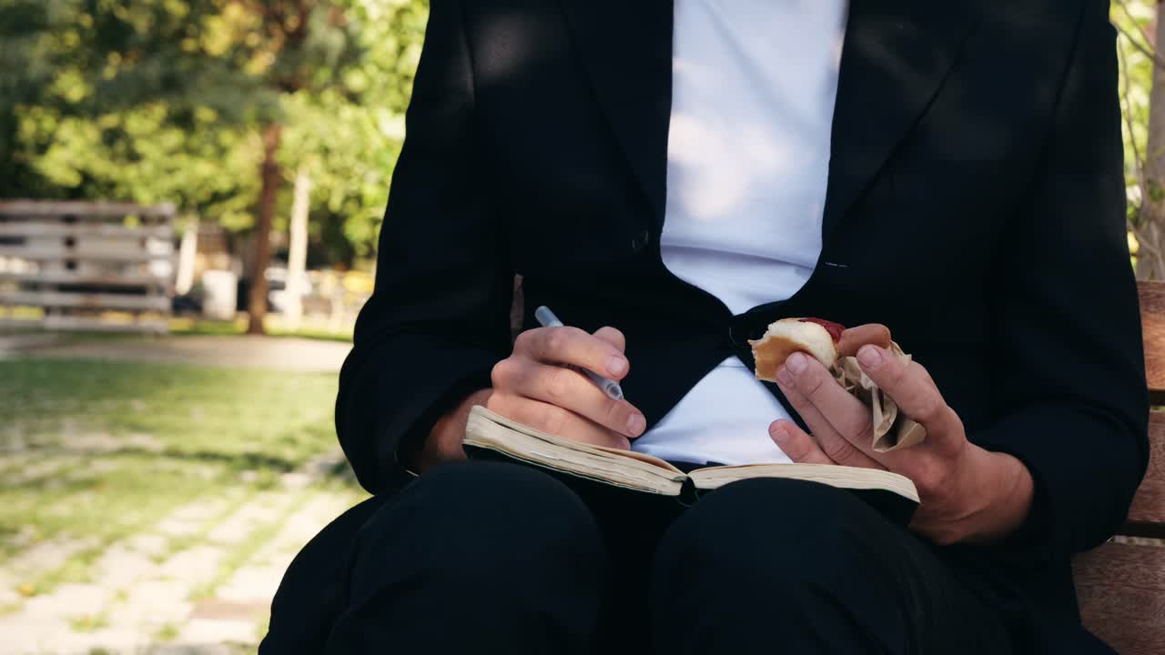 Man in suit writing in notebook while eating sandwich in the park