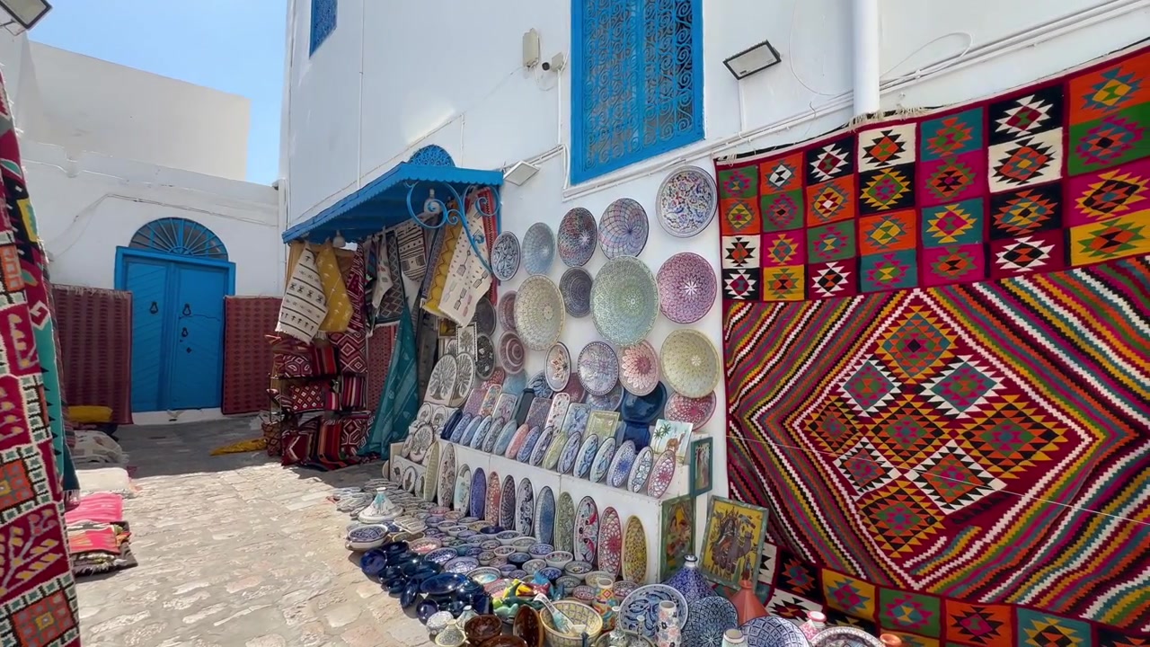 Des assiettes en céramique, des bols et des tapis tissés bordent une ruelle en pierre tranquille à Sidi Bou Saïd sous une lumière vive de midi, mettant en valeur l'artisanat local et l'ambiance du marché. Tunisie
