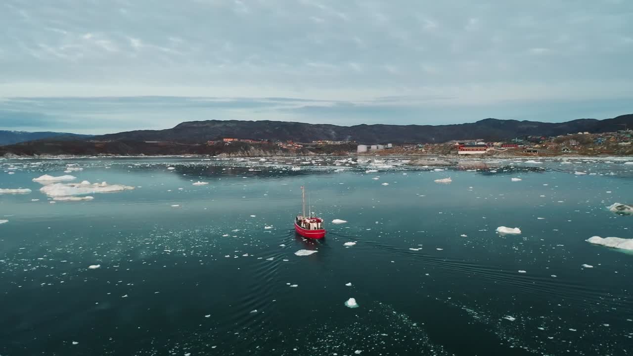 A red fishing boat sails through icy Arctic waters near Greenland, surrounded by floating icebergs and snow-capped mountains in the distance, captured by a drone on a calm, cold day