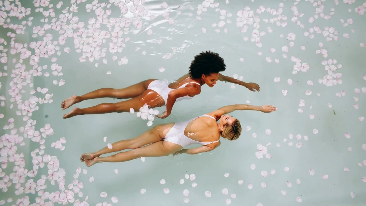 Floating women starting aligned, stroking, staying aligned in pool, wearing white suits amid petals