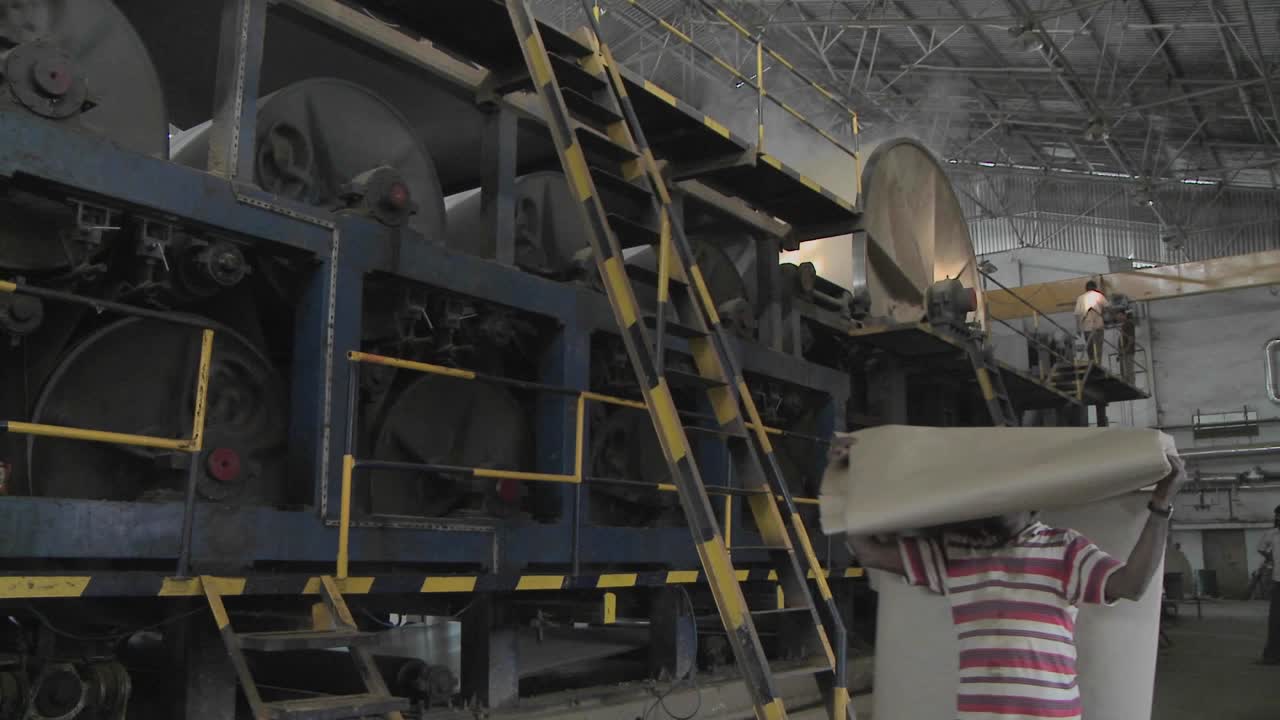 A man carries a large pile of paper over his head through a manufacturing plant