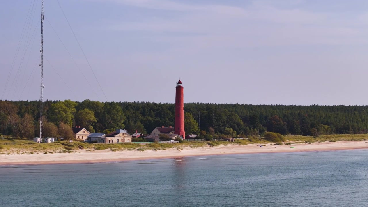 Ripresa aerea panoramica in ampia orbita del faro rosso di Akmenrags sulla costa di Kurzeme in Lettonia, circondato dal mare e da spiagge sabbiose.