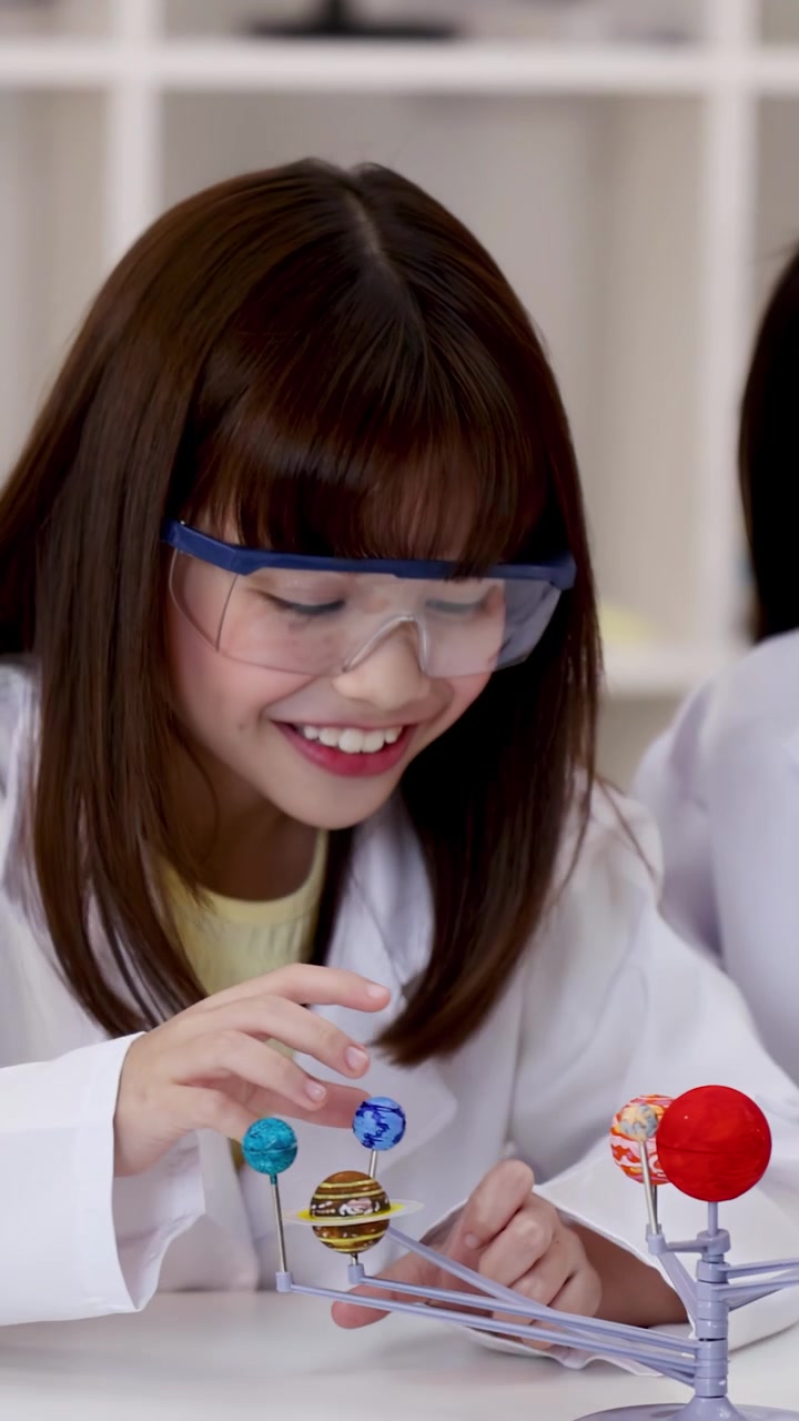 Smiling student wearing safety goggles interacts with a planetary model in a bright classroom.