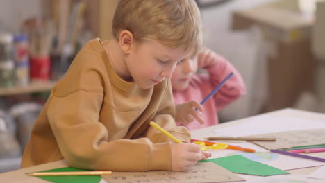 blonde little girl and blond kid drawing on a paper sitting at a table where are sign with environmental quotes in a craft workshop