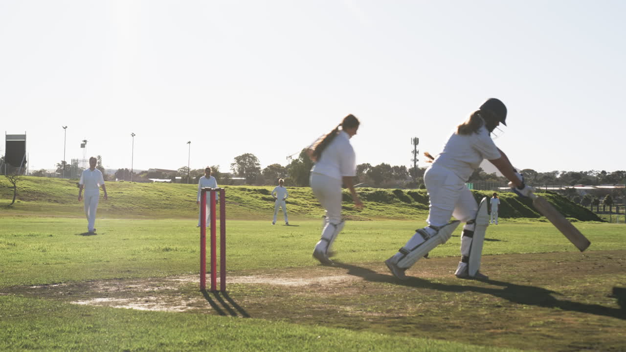 Playing cricket on sunny day, female players batting and fielding on grass field