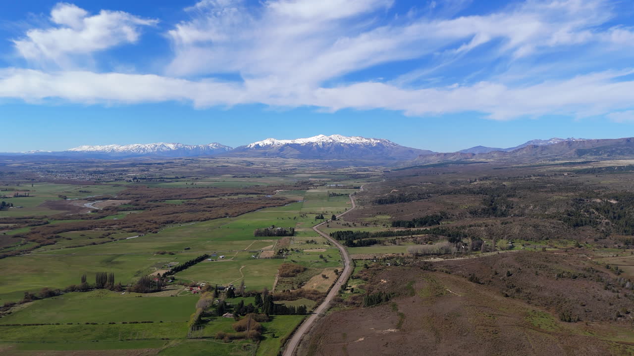 High altitude drone flyover shot in green and brown fields, with deep blue skies, some clouds and snowed mountains in back. Shot on 4K-60fps