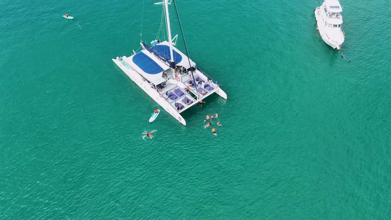 푸켓의 푸르키즈색 물에서 카타마란 주위에 헤엄치는 사람들의 항공 사진, 활기차고 이 많은 휴가 장면을 보여줍니다.