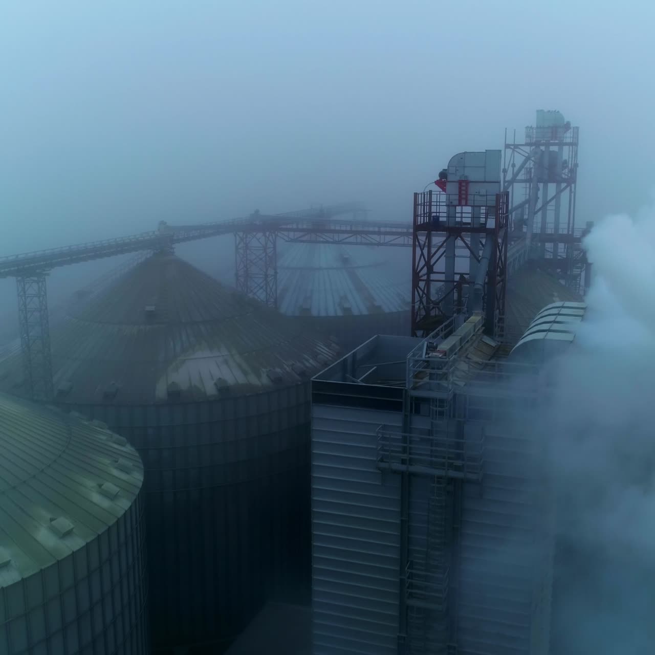 Tops of the elevator tanks hardly seen in grey fog and thick white smoke coming from pipes. Aerial view on the modern granary complex plant
