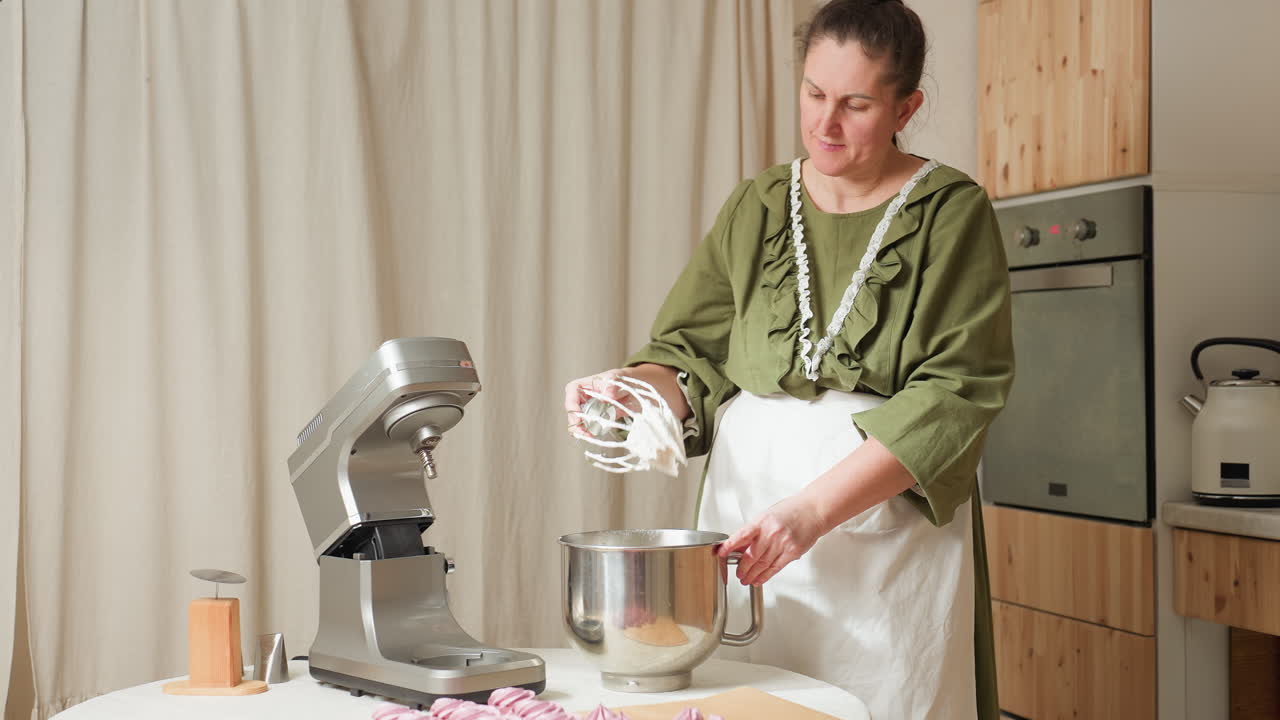 Baker in green dress and apron removes dough from electric mixer in kitchen, beating sticky mixture against mixing bowl to clean beaters, surrounded by baking tools and oven in warm indoor