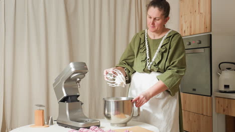 baker in green dress and apron removes dough from electric mixer in kitchen, beating sticky mixture against mixing bowl to clean beaters, surrounded by baking tools and oven in warm indoor