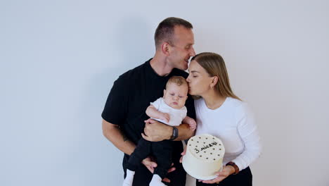 beautiful caucasian family celebrating three months of their newborn. mom holds a festive cake and kisses her child. man kisses his wife on the head.
