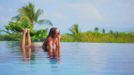 Stunning-girl-lying-on-her-belly-in-shallow-water-inside-infinity-pool-enjoying-gorgeous-tropical-scenery,-daytime-slow-motion