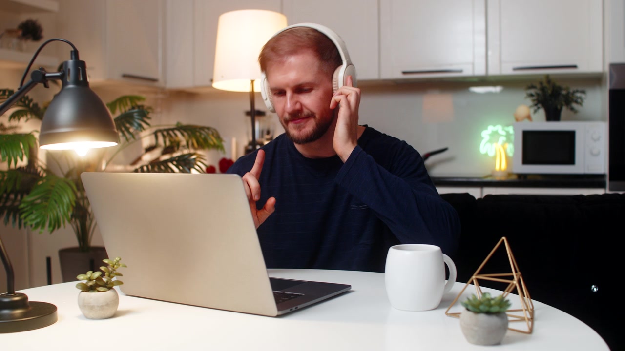 Ung mandlig freelancer med fokus på bærbar computer, der har trådløse hovedtelefoner på, drikker te og arbejder om natten indendørs