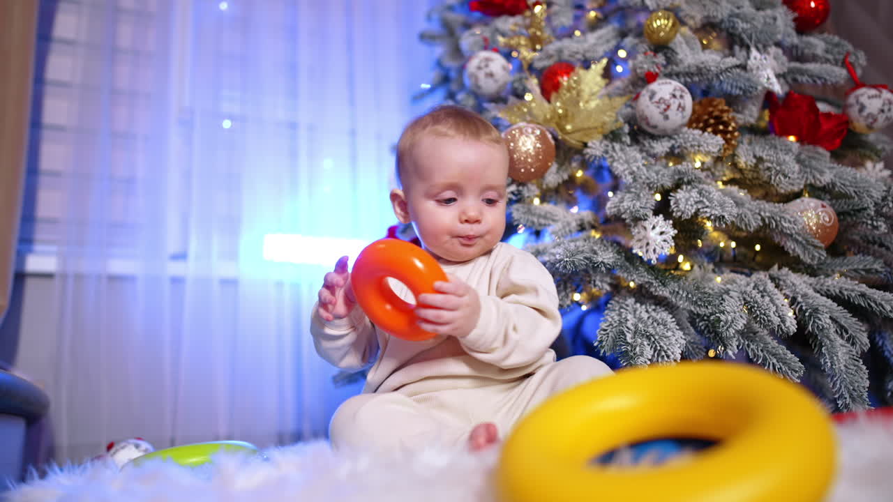 Beautiful baby in white clothes sits near the decorated Christmas tree. Happy child plays with toys trying to put them to his mouth as he's teething. Low angle view.