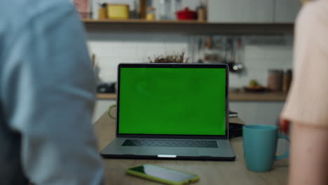 una pareja viendo una computadora portátil de pantalla verde sentada en la mesa de la cocina con un café de cerca