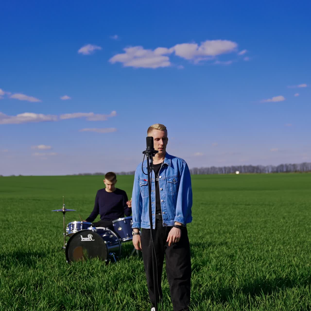 Lead singer approaches microphone stand slowly. Blond guy singing outdoors. Musicians playing music at backdrop in the green field in blur