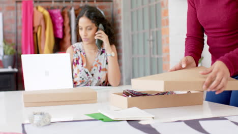 diverse female fashion designers using laptop, on phone and packing order in studio, slow motion