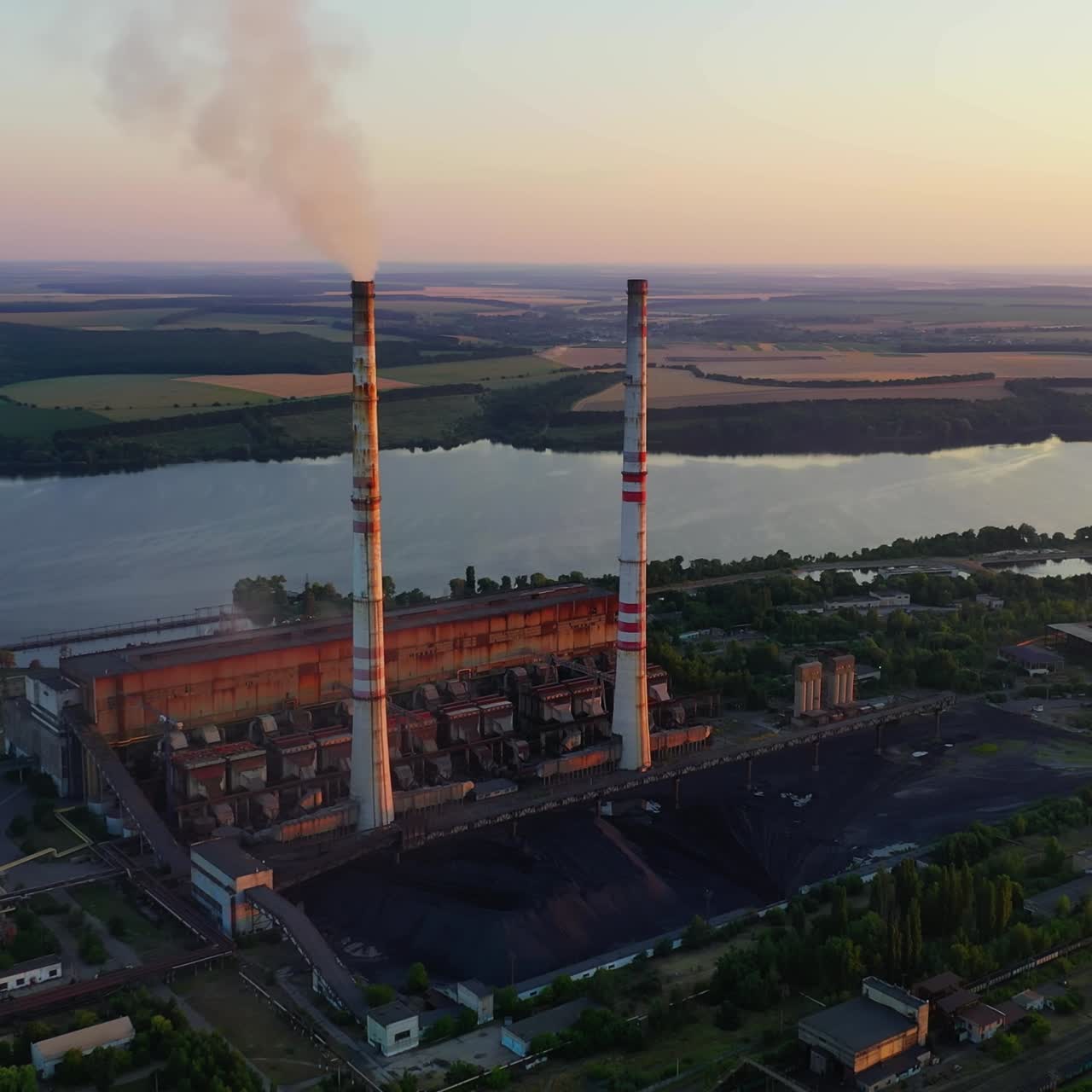 Industrial enterprise on nature background. Chemical plant with pipes near the river. Dirty smoke pours into the air from factory. Environmental pollution. Aerial view
