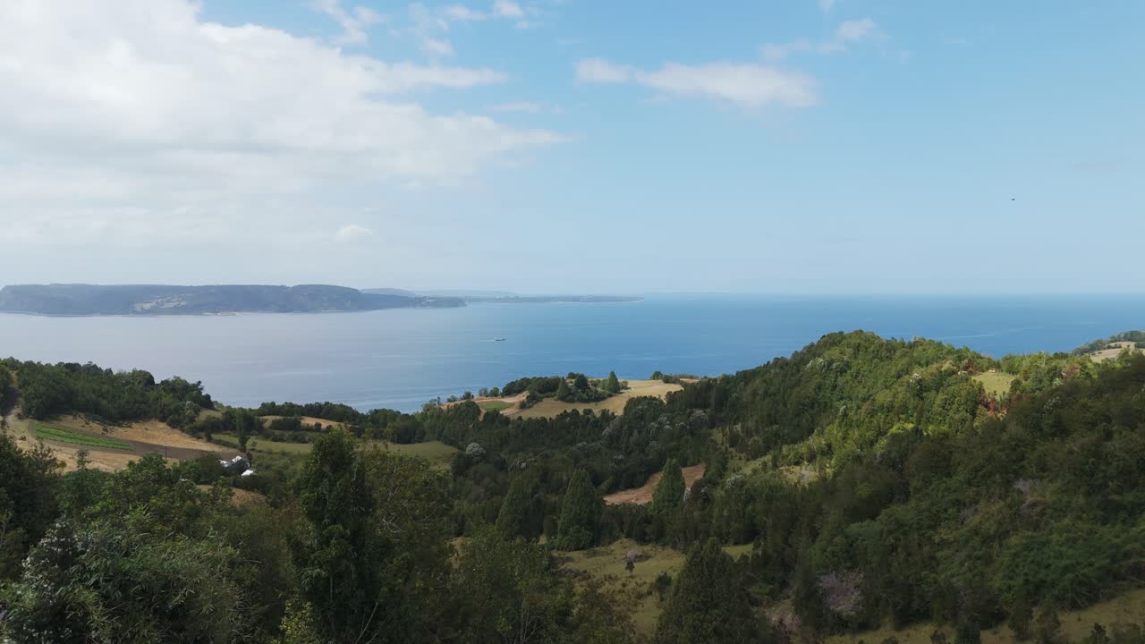 Closed parasols shading a wooden bench overlooking Reloncavi Sound and the green hills of Lemuy Island, Chile, on a beautiful sunny day. Aerial dolly forward
