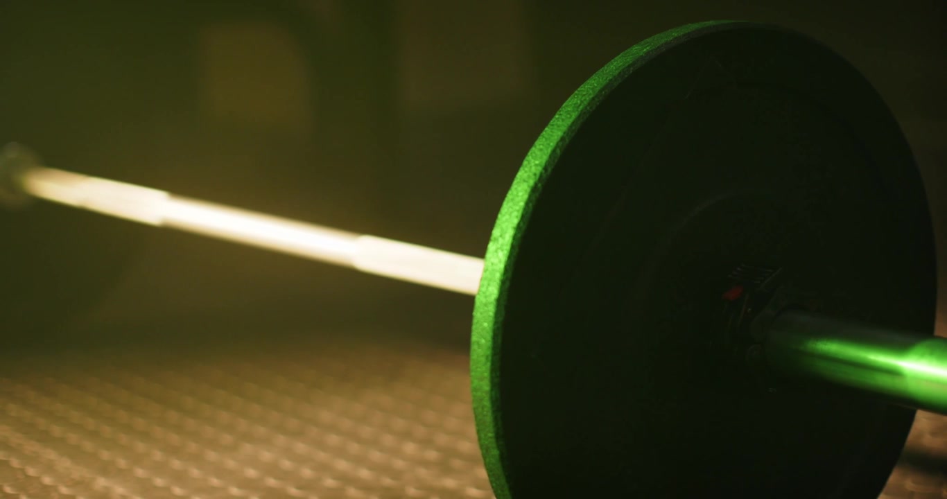 La telecamera si sta spostando, la luce si sta schiarendo, una piastra verde e un bilanciere stanno rivelando il pavimento in gomma della palestra.