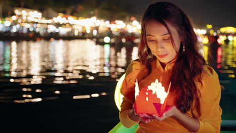 vietnamese woman in yellow dress prays with paper candle on river boat at night in hoi an, vietnam in slow motion