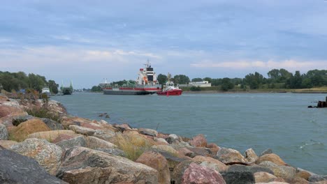 großes graues frachtschiff, das an einem ruhigen, sonnigen sommertag in den hafen von liepaja einläuft, steinpier im vordergrund, oskara kalpaka-drehbrücke im hintergrund, plätschernde wellen, weitwinkelaufnahme