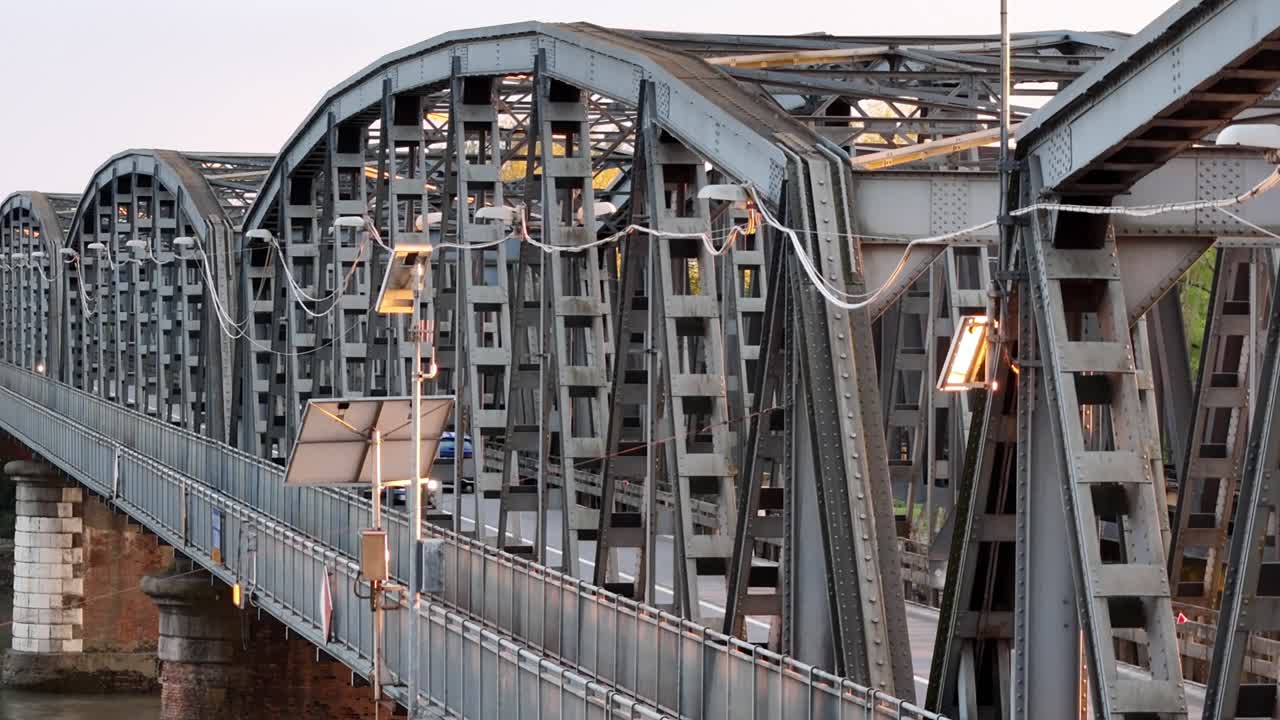 Drone ascends steadily along the iron bridge near Cremona, capturing the structural design and vehicles crossing at sunset, with warm lighting on steel beams and urban-rural elements in the background