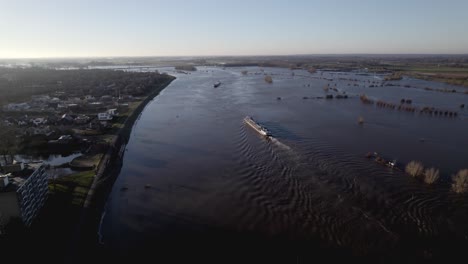 Luftanflug-Von-Großen-Binnenfrachtschiffen,-Die-Bei-Hohem-Wasserstand-Wellen-Auf-Dem-Fluss-Ijssel-Hinterlassen,-Mit-überfluteten-Auen-Der-Turmstadt-Zutphen-In-Den-Niederlanden