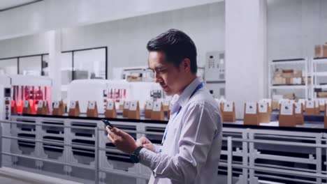 side view of an asian male professional worker standing with his smartphone with conveyor for packaging coffee beans in bags at coffee factory, checking on his smartphone with meditation