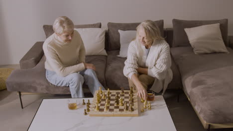 two senior women playing chess sitting on sofa at home 4