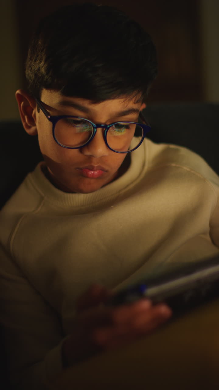 Vertical Video Close Up Of Young Boy Sitting On Sofa At Home Playing Game Or Streaming Onto Handheld Gaming Device At Night