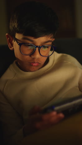 vertical video close up of young boy sitting on sofa at home playing game or streaming onto handheld gaming device at night