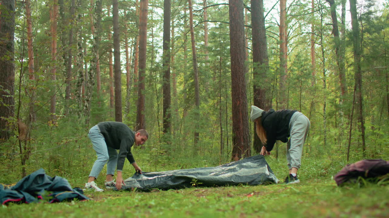 Man and woman arrange camping equipment on forest ground with bag on floor among tall pine trees and green undergrowth, coordinating gear setup during peaceful outdoor adventure morning