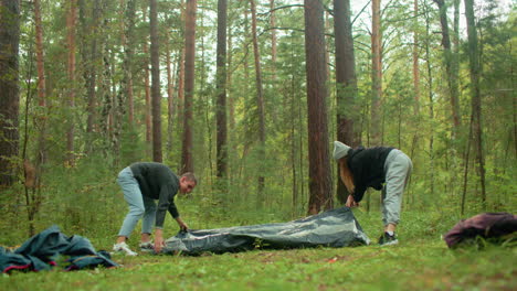 man and woman arrange camping equipment on forest ground with bag on floor among tall pine trees and green undergrowth, coordinating gear setup during peaceful outdoor adventure morning
