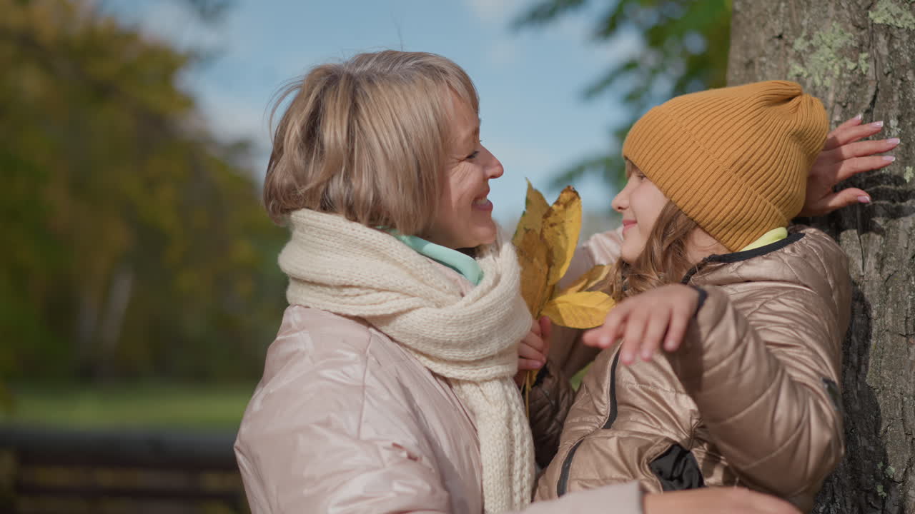 little girl leans against tree trunk in quiet moment wearing warm coat and mustard hat while her mother gently offers bundle of autumn leaves and wraps her in warm loving hug with smile