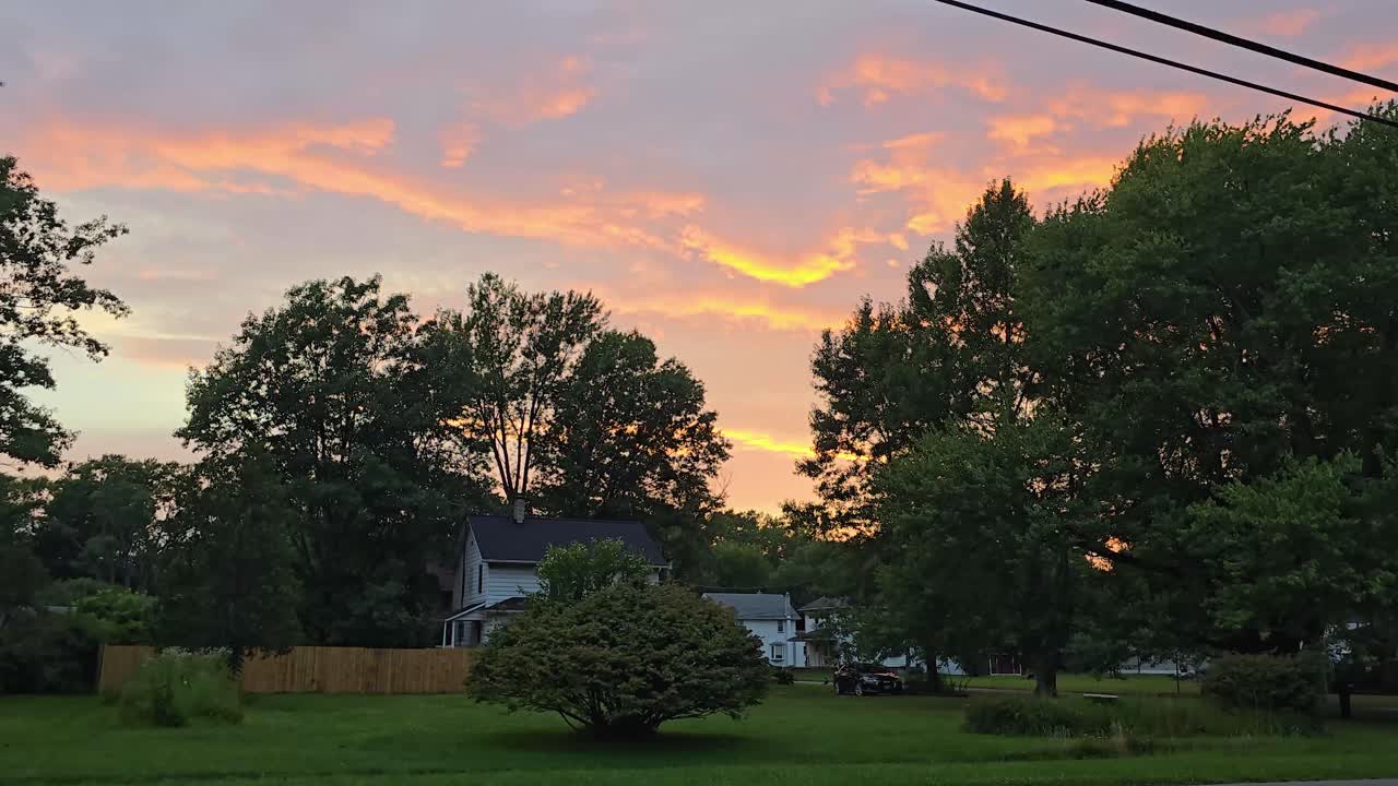 산란색과 분홍색의 색조로 활기찬 일몰이 평화로운 교외 동네 위의 하늘을 비추고, 나무와 지붕으로 둘러싸여 있습니다.