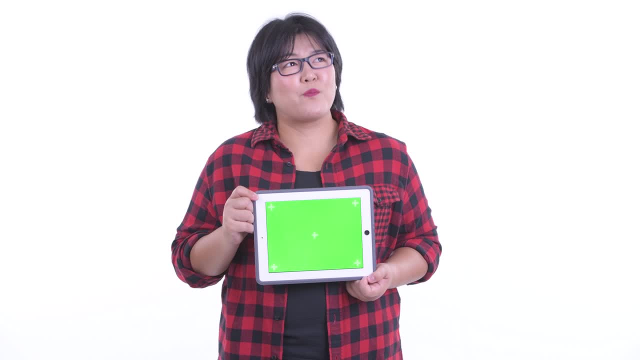 색 스튜디오 배경에 고립되어 미소 짓고 생각하면서 복사 공간을 가진 디지털 태블릿을 들고 있는 아름다운 과체중 아시아 여성의 초상화